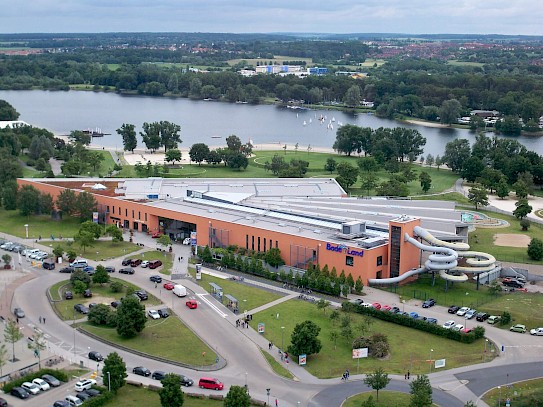 Das orangefarbene BadeLand Wolfsburg mit Parkplatz aus der Vogelperspektive und dem Allerpark im Hintergrund