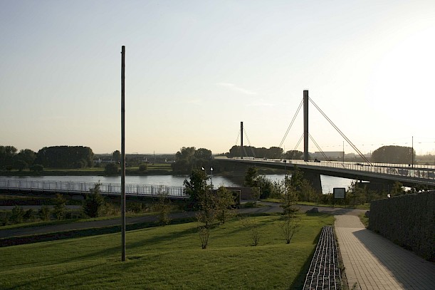 Grünflächiges Ufer mit Spazierweg und Blick auf das Rheinufer. In der Ferne eine Brücke und wolkenloser Himmel.