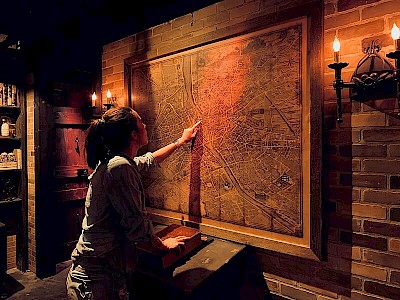 Dunkler Raum, der nur von Kerzenlicht erhellt wird. Eine Frau steht vor einer Ziegelwand, an der eine Landkarte hängt.