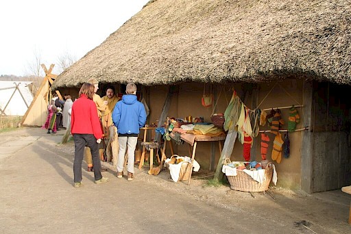 Museumsbesucher:innen vor einer Lehmhütte mit Strohdach, wo ein Bewohner Kleidung verkauft.